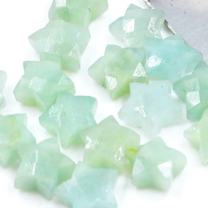 May include: A collection of light green star-shaped gemstone beads. The beads are scattered on a white surface and spilling out of a metal scoop.