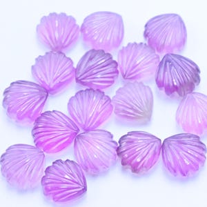May include: A collection of small, translucent, shell-shaped beads in shades of purple. Each bead has a detailed, fan-like pattern. The beads are scattered on a white surface, creating a delicate and artistic arrangement.