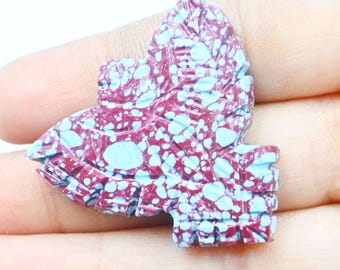 May include: A carved stone butterfly with a maroon and turquoise speckled pattern. The butterfly has detailed wing and body features. The stone is held in a hand against a white background.