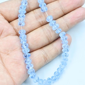 Può includere: Una collana di perline traslucide azzurre. Ogni perlina presenta un caratteristico motivo a coste. Le perline sono infilate su un sottile filo bianco. La collana è tenuta nella mano di una persona su sfondo bianco.