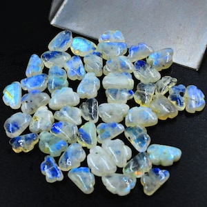 May include: A collection of white moonstone gemstone beads with iridescent blue and green hues. The beads are shaped like irregular clouds and are scattered on a black surface.