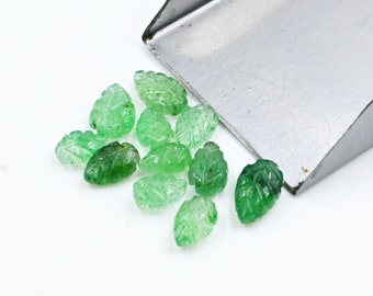 Puede incluir: Un grupo de piedras preciosas verdes en forma de hoja, algunas con un tono verde más oscuro, están esparcidas sobre una superficie blanca. Las piedras preciosas están talladas con intrincados detalles, que se asemejan a hojas. Una pala de metal es parcialmente visible en la esquina superior derecha de la imagen.