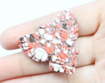 May include: A carved stone leaf-shaped pendant with a mosaic pattern of red, white, and black fragments. The pendant is held in a hand, showcasing its intricate design and color variations. The leaf has a detailed, textured surface.