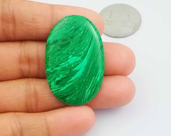 May include: A green oval malachite cabochon gemstone with swirling patterns. The stone is being held in a hand for size comparison.