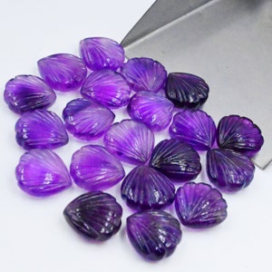 May include: A collection of 18 purple amethyst gemstone flower petals, each carved with a detailed shell-like design. The petals are arranged on a silver surface.