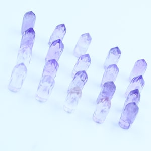 May include: A collection of small, purple amethyst crystal points arranged in rows on a white background.