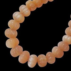 Puede incluir: Una hebra de cuentas de piedras preciosas de color melocotón con una superficie tallada y texturizada. Las cuentas son redondas y tienen una apariencia ligeramente translúcida.