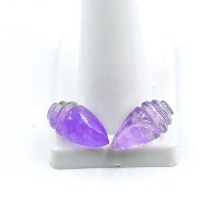 May include: Two carved purple gemstone pendants, shaped like stylized shells. The pendants are a vibrant shade of lavender and are set against a white background. The shell design features detailed spiral carvings.
