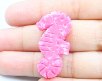May include: A pink seahorse figurine with white marbling. The seahorse is a detailed, three-dimensional design, with visible ridges and a curled tail. The figurine is held in a hand, showcasing its size and shape.