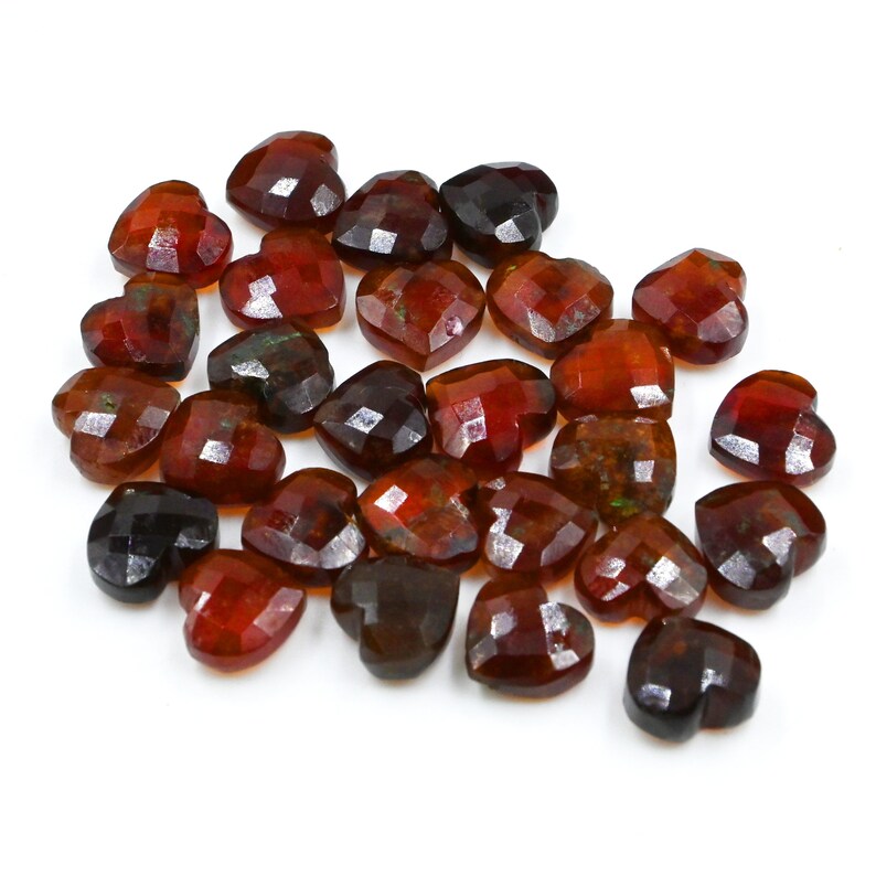 May include: A collection of 25 faceted heart-shaped gemstones in shades of brown, red, and green. The stones are arranged in a loose pile on a white background.