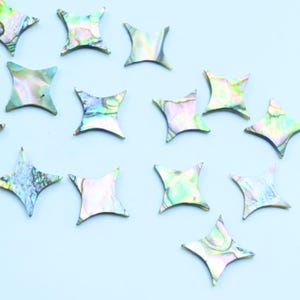 Könnte beinhalten: Iridescent schimmernde, sternförmige Muschelteile auf hellblauem Grund. Die Teile haben einen natürlichen, perlmuttartigen Glanz mit grünen, rosa und blauen Farbtönen. Ideal für Bastelarbeiten und Schmuckherstellung.