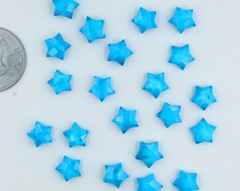 May include: Twenty-two turquoise blue star-shaped cabochons, each with a slightly different shape and size, are scattered on a white surface.