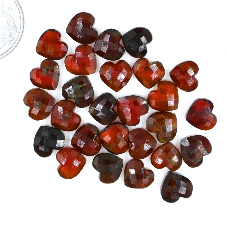 May include: A collection of 20 heart-shaped, faceted gemstones in shades of red, brown, and black. The stones are arranged in a loose pile on a white background.