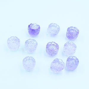 May include: Ten purple amethyst flower-shaped beads with intricate carvings. The beads are smooth and polished, with a light purple hue.
