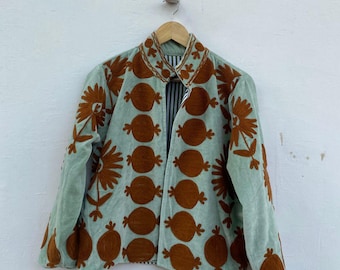 May include: A light green jacket with brown floral embroidery. The jacket is hanging on a wooden hanger against a white wall. The embroidery features a repeating pattern of circular shapes and flowers.