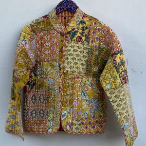 Könnte beinhalten: Gesteppte Jacke mit Patchwork-Design in Gelb-, Grün- und Rosatönen. Die Jacke mit floralem Muster hängt an einem Kleiderbügel. Sie hat eine Knopfleiste und zwei Taschen. Maße in Zentimetern.