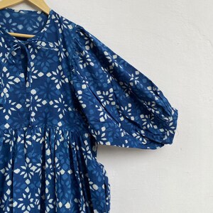 May include: A blue dress with a white floral pattern is displayed on a wooden hanger. The dress features short sleeves, a gathered waist, and a tie at the neckline. The fabric has a textured appearance.