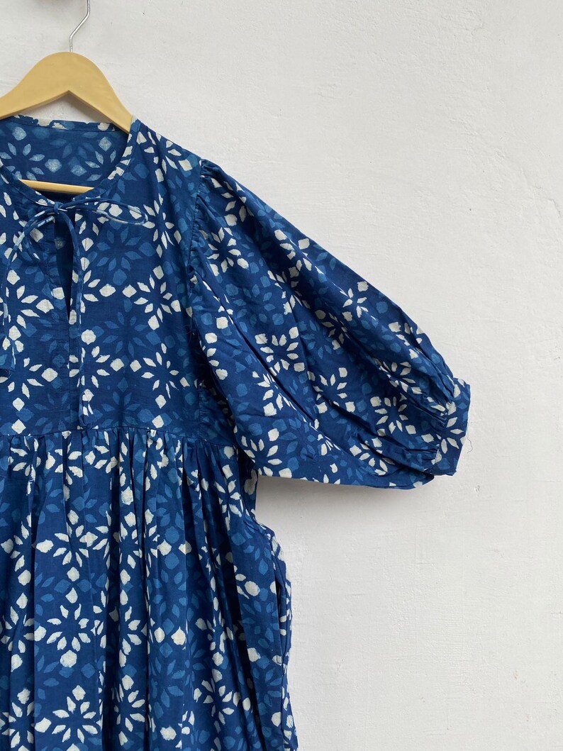 May include: A blue dress with a white floral pattern is displayed on a wooden hanger. The dress features short sleeves, a gathered waist, and a tie at the neckline. The fabric has a textured appearance.