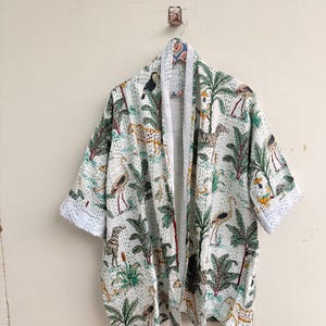 Op de afbeelding: Een wit kimonojack met een herhalende jungleprint van groene palmbomen, kleurrijke vogels en gevlekte luipaarden. Het kledingstuk heeft een open voorzijde, wijde driekwartmouwen met omgeslagen boorden en zichtbare witte stiksels.