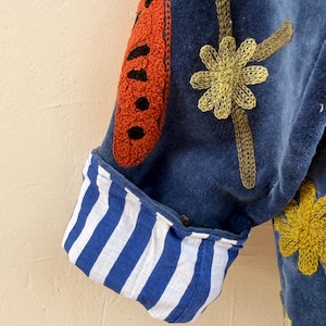 May include: A blue velvet jacket sleeve featuring a rolled cuff with a blue and white vertical stripe pattern. The jacket is decorated with embroidered orange and black butterfly wing motifs and yellow flower designs on the blue velvet fabric.