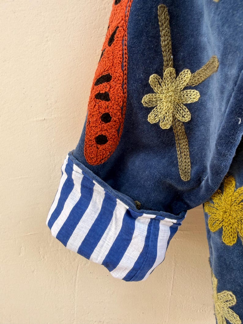 May include: A blue velvet jacket sleeve featuring a rolled cuff with a blue and white vertical stripe pattern. The jacket is decorated with embroidered orange and black butterfly wing motifs and yellow flower designs on the blue velvet fabric.