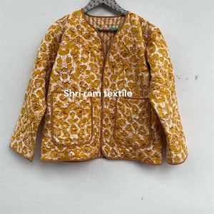 Op de afbeelding: Een gewatteerde jas met een bloemenpatroon in geel en wit, afgewerkt met bruine randen. De jas heeft twee voorzakken en de tekst "Shri ram textile". Het is een lichtgewicht kledingstuk, hangend aan een decoratieve hanger.