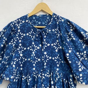 May include: A blue dress with a white floral pattern is displayed on a wooden hanger. The dress features a round neckline with a tie, short puffed sleeves, and a gathered skirt. The fabric has a textured appearance.