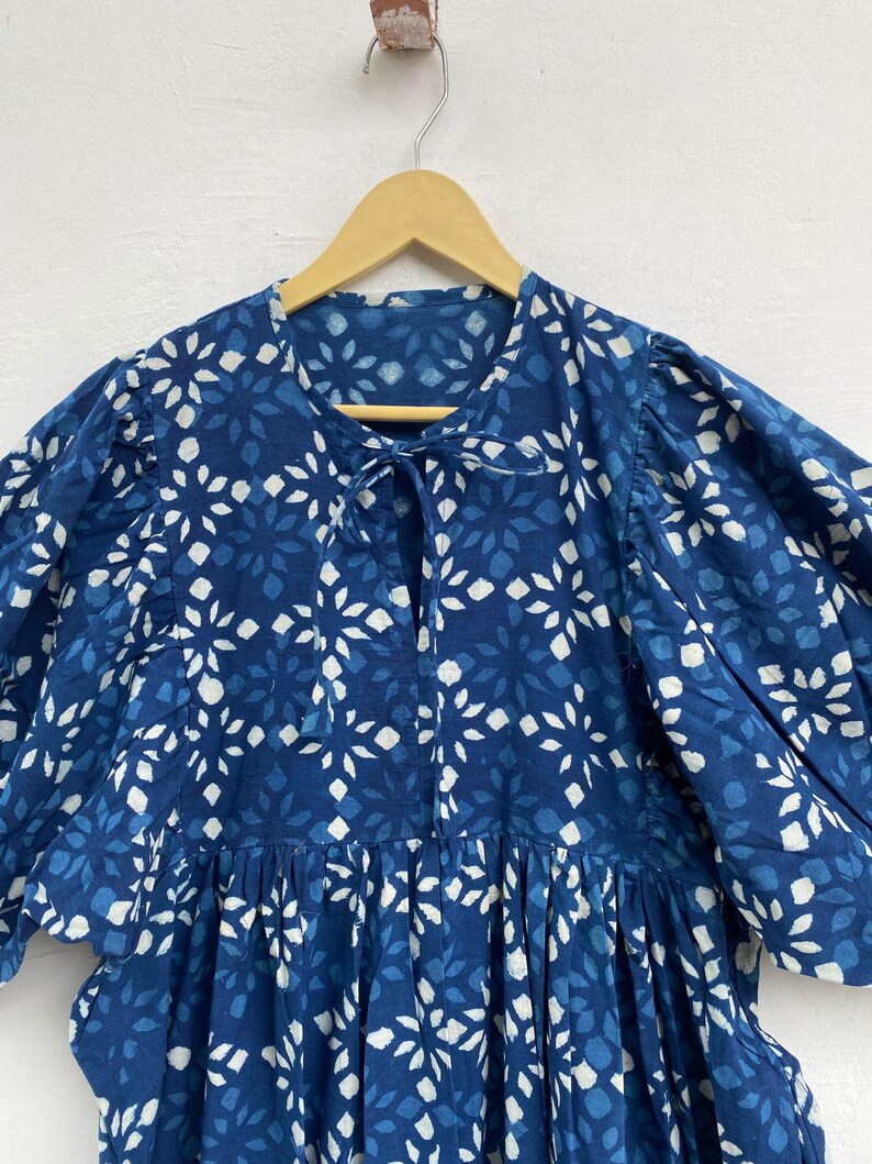 May include: A blue dress with a white floral pattern is displayed on a wooden hanger. The dress features a round neckline with a tie, short puffed sleeves, and a gathered skirt. The fabric has a textured appearance.