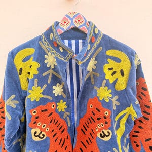 May include: A blue jacket featuring vibrant orange tiger embroidery and yellow floral motifs. The garment has a high collar, long sleeves, and a white and blue vertical striped lining visible at the front opening. The fabric appears to be velvet.