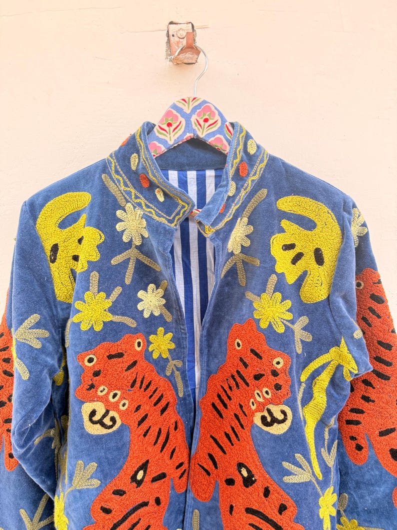 May include: A blue jacket featuring vibrant orange tiger embroidery and yellow floral motifs. The garment has a high collar, long sleeves, and a white and blue vertical striped lining visible at the front opening. The fabric appears to be velvet.