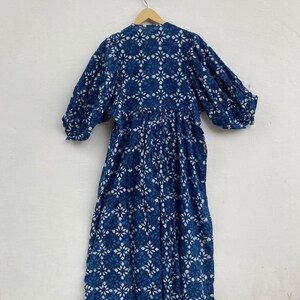May include: A long, flowing dress in a deep indigo hue, adorned with a repeating white floral pattern. The dress features long, voluminous sleeves and a gathered waist, hanging from a wooden hanger against a white wall. The fabric appears lightweight and breathable.