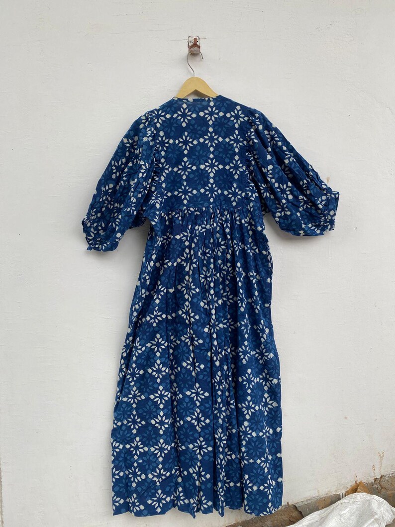 May include: A long, flowing dress in a deep indigo hue, adorned with a repeating white floral pattern. The dress features long, voluminous sleeves and a gathered waist, hanging from a wooden hanger against a white wall. The fabric appears lightweight and breathable.