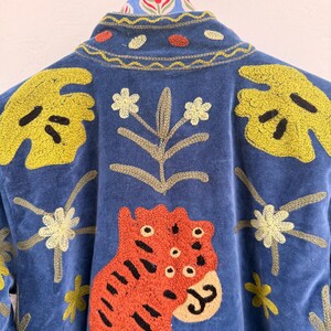 May include: A blue jacket featuring vibrant embroidery of a large orange tiger, yellow monstera leaves, and small white flowers. The collar displays a yellow wavy stitch pattern with small orange and yellow circular accents, all on a blue fabric.