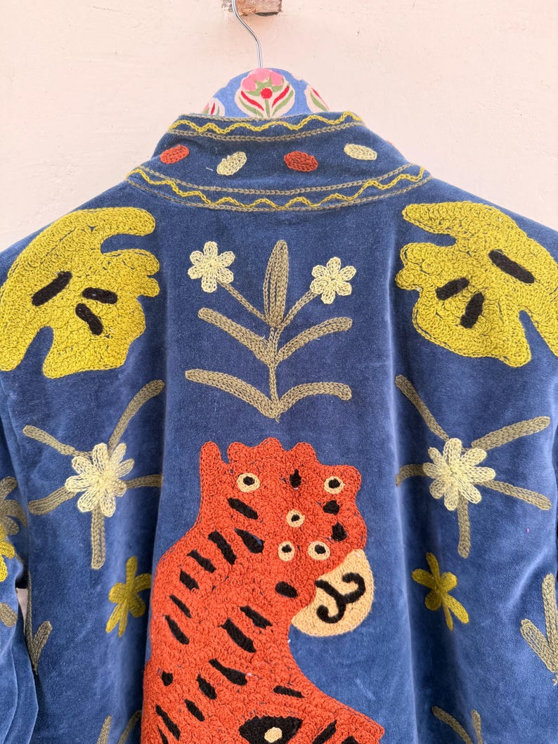 May include: A blue jacket featuring vibrant embroidery of a large orange tiger, yellow monstera leaves, and small white flowers. The collar displays a yellow wavy stitch pattern with small orange and yellow circular accents, all on a blue fabric.