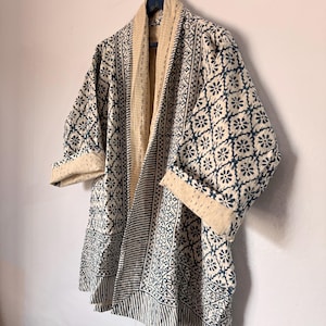 Puede incluir: Una chaqueta estilo kimono con estampado azul y blanco con interior color crema. La chaqueta presenta un diseño floral y geométrico, con mangas enrolladas y un cuello color crema en contraste. La chaqueta está colgada en una pared.