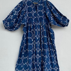 May include: A long, flowing dress in a deep indigo blue, adorned with a repeating white floral pattern. The dress features a round neckline with a tie, three-quarter sleeves, and a gathered waist, creating a relaxed silhouette. The dress is hanging on a wooden hanger.