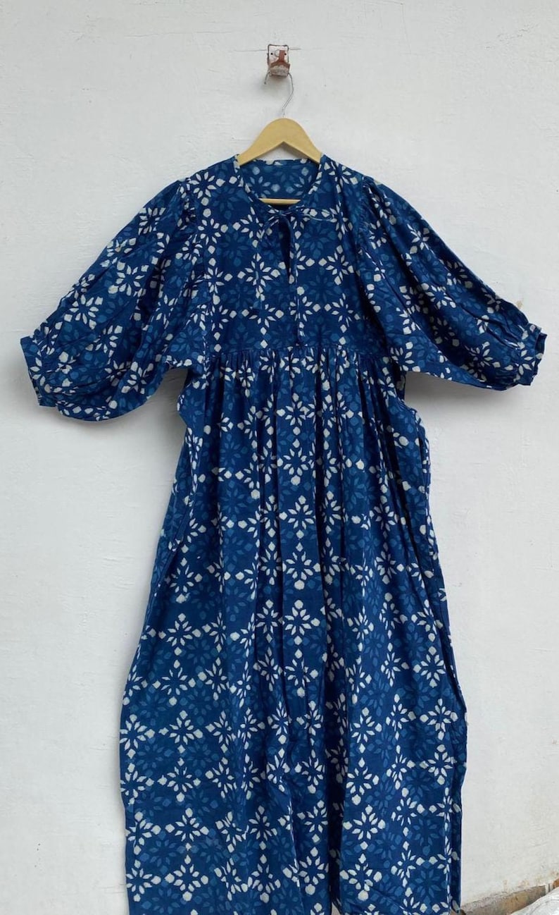 May include: A long, flowing dress in a deep indigo blue, adorned with a repeating white floral pattern. The dress features a round neckline with a tie, three-quarter sleeves, and a gathered waist, creating a relaxed silhouette. The dress is hanging on a wooden hanger.