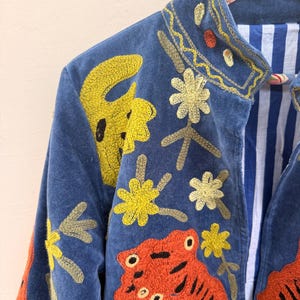 May include: A blue velvet jacket featuring vibrant embroidery of orange tigers and yellow flowers. The garment has a stand-up collar and an interior lining with blue and white vertical stripes. The jacket is displayed on a patterned hanger.