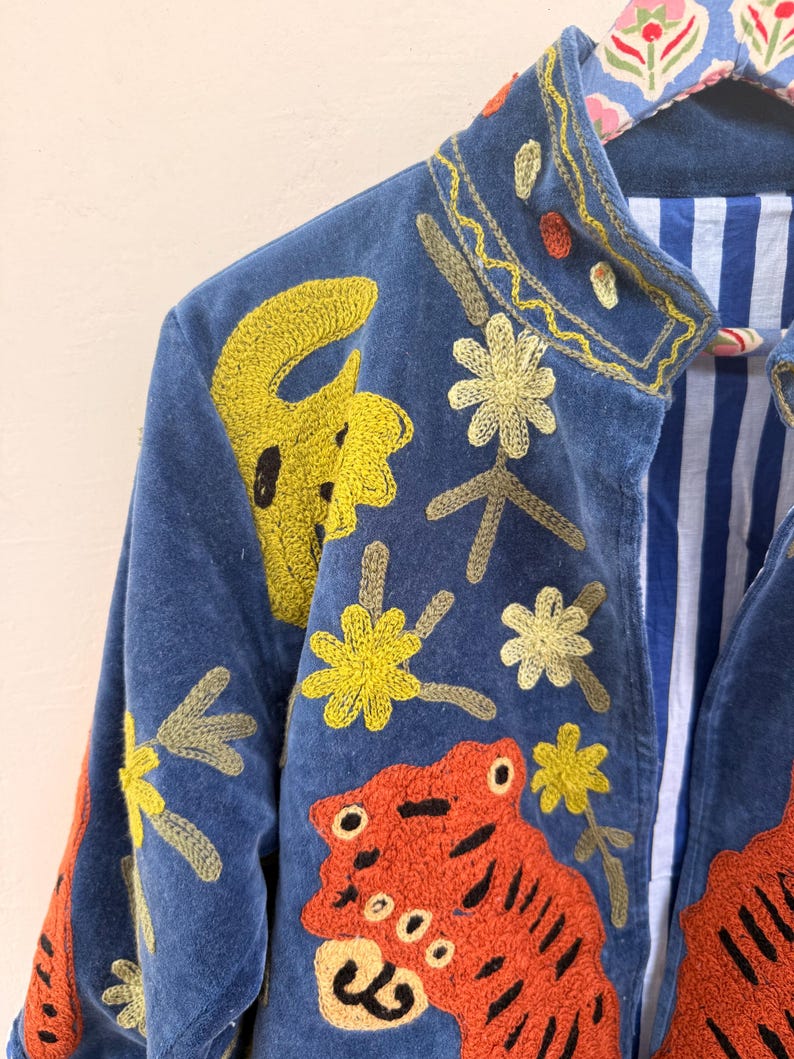 May include: A blue velvet jacket featuring vibrant embroidery of orange tigers and yellow flowers. The garment has a stand-up collar and an interior lining with blue and white vertical stripes. The jacket is displayed on a patterned hanger.