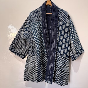 May include: A navy blue and white patterned jacket with a shawl collar, hanging on a wooden hanger. The jacket features a patchwork design with various geometric and floral patterns. The sleeves are short, and the jacket is made of a quilted fabric.