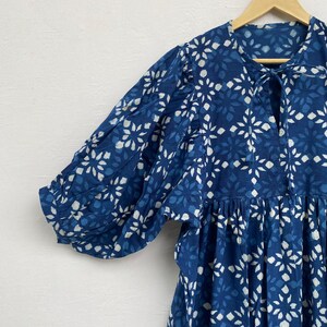May include: A blue dress with a white floral pattern, hanging on a wooden hanger. The dress features short, puffy sleeves and a tie at the neckline. The fabric has a textured appearance.