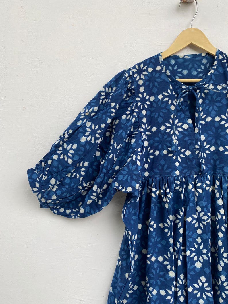 May include: A blue dress with a white floral pattern, hanging on a wooden hanger. The dress features short, puffy sleeves and a tie at the neckline. The fabric has a textured appearance.