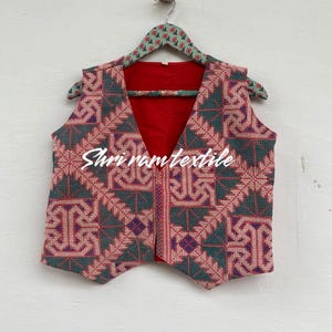 Puede incluir: Un chaleco sin mangas con cuello en V, con un patrón geométrico en tonos rojos, verdes y morados. El chaleco tiene un forro rojo y está colgado de una percha con estampado floral. El texto "Shri ram textile" es visible.
