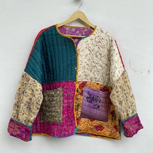 Op de afbeelding: Een patchwork jasje met diverse patronen en kleuren, waaronder teal, crème, roze en bloemenprints. De jas heeft twee zakken en is gewatteerd. Het hangt aan een houten hanger.