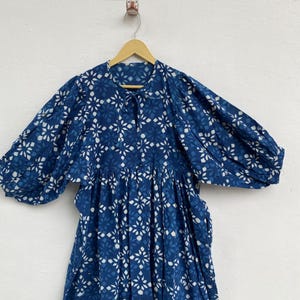 May include: A blue dress with a white floral pattern is displayed on a wooden hanger. The dress has long, puffy sleeves and a gathered waist. The fabric appears to be lightweight, and the dress is hanging against a white wall.
