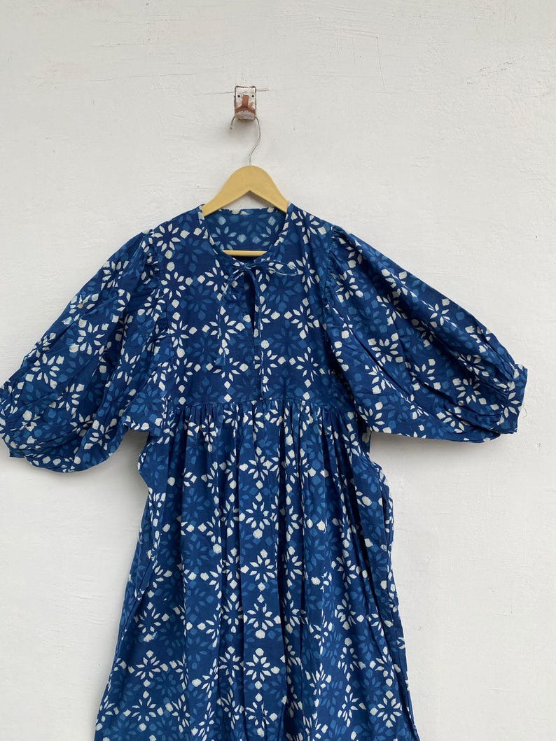 May include: A blue dress with a white floral pattern is displayed on a wooden hanger. The dress has long, puffy sleeves and a gathered waist. The fabric appears to be lightweight, and the dress is hanging against a white wall.