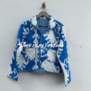 Puede incluir: Chaqueta acolchada con estampado ikat azul y blanco. La chaqueta tiene dos bolsillos delanteros y un cuello. El forro interior es de rayas azules y blancas. La chaqueta está colgada de una percha estampada.