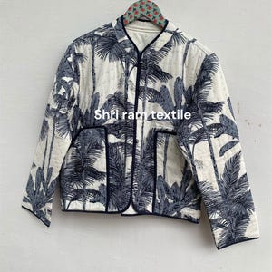 Puede incluir: Chaqueta acolchada con fondo blanco y un estampado repetido de palmeras azul oscuro. La chaqueta tiene cuello en V y está ribeteada en azul oscuro. El texto "Shri ram textile" está impreso en la chaqueta.