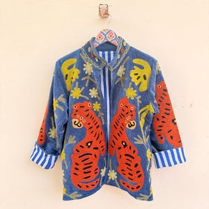May include: A blue jacket featuring vibrant orange tiger embroidery on the front panels, accented with yellow floral motifs. The garment has a high collar, long sleeves with blue and white striped cuffs, and a matching blue and white striped interior lining.