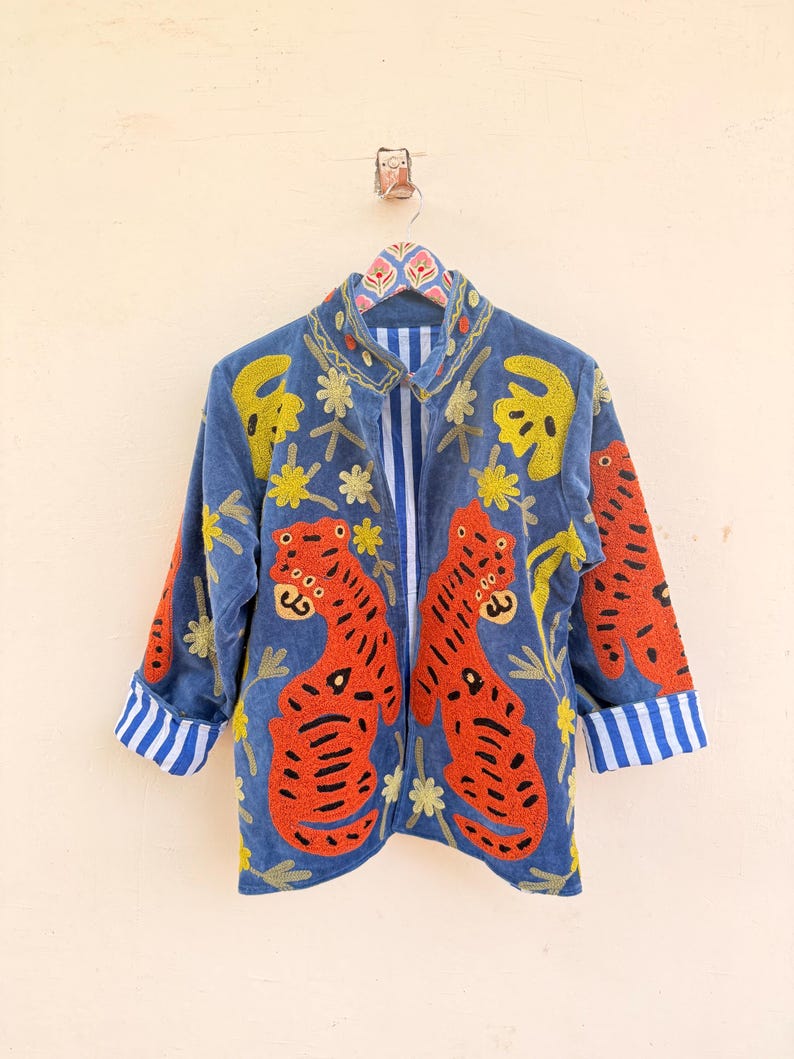 May include: A blue jacket featuring vibrant orange tiger embroidery on the front panels, accented with yellow floral motifs. The garment has a high collar, long sleeves with blue and white striped cuffs, and a matching blue and white striped interior lining.